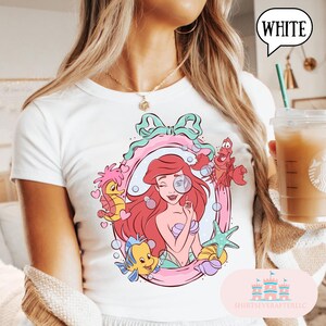 May include: White t-shirt featuring a colorful graphic of Ariel from The Little Mermaid, framed by a pink and teal border. The design includes seahorses, a starfish, and other characters. The word "WHITE" is in a speech bubble.