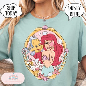 May include: Dusty blue t-shirt featuring a colorful illustration of Ariel and Flounder from The Little Mermaid. The graphic is framed with a gold border and pink accents. The shirt has the text "Ship Today" and "Dusty Blue".