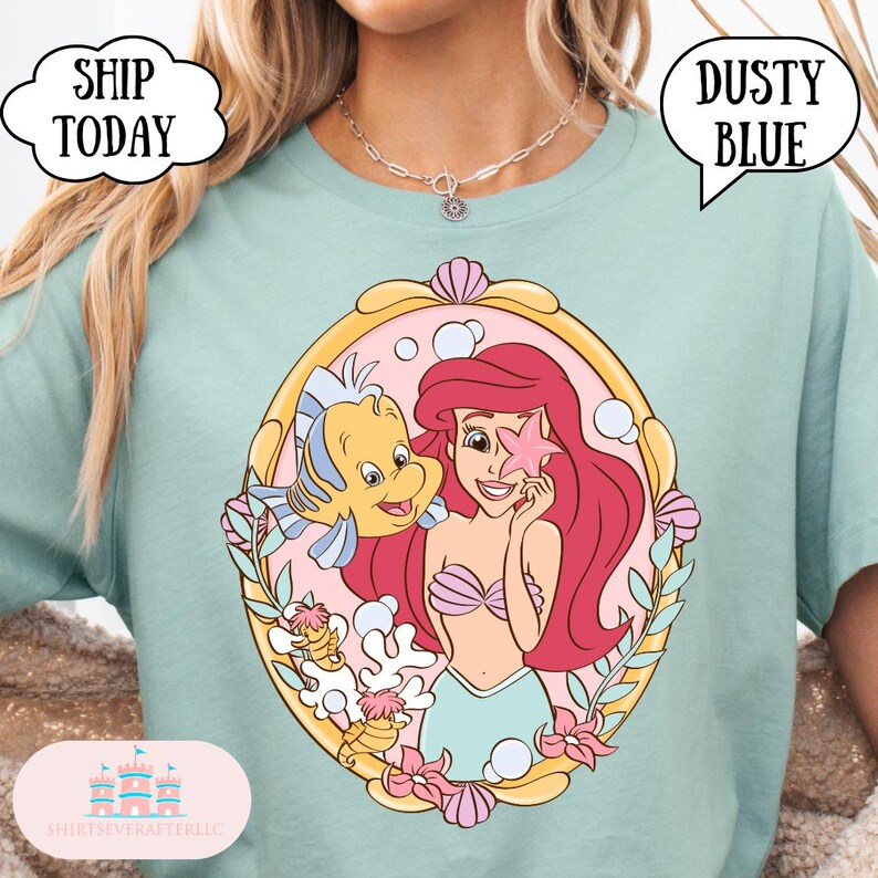 May include: Dusty blue t-shirt featuring a colorful illustration of Ariel and Flounder from The Little Mermaid. The graphic is framed with a gold border and pink accents. The shirt has the text "Ship Today" and "Dusty Blue".