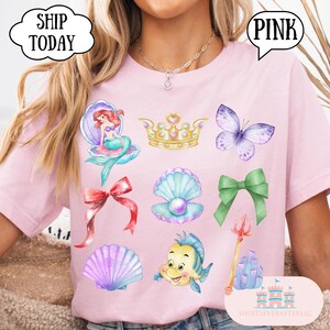 Może przedstawiać: R&oacute;żowa koszulka z akwarelowymi ilustracjami syreny, korony, motyla, kokardy, muszli i ryby. Koszulka ma napis "PINK" w dymku i "SHIP TODAY" w chmurce.