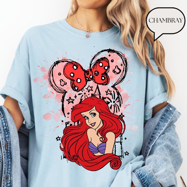 May include: Light blue t-shirt featuring a graphic of Ariel from The Little Mermaid, with a red bow and Mickey Mouse ears design. The design includes red and pink accents, stars, and a black outline. The shirt has rolled-up sleeves.