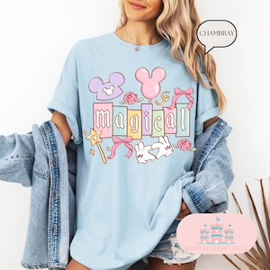 Op de afbeelding: Lichtblauw T-shirt met het woord "magical" in kleurrijke blokletters. Het ontwerp bevat een Mickey Mouse-hoofd, een roze ballon, een toverstaf, strikken, rozen en een konijn. Het shirt maakt deel uit van de collectie "Shirts Ever After LLC".