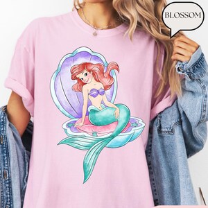 May include: Pale pink t-shirt with a watercolour illustration of a mermaid in a seashell. The mermaid has red hair, a purple seashell bra, and a teal tail. The seashell is purple and blue. The word "BLOSSOM" is in a speech bubble.