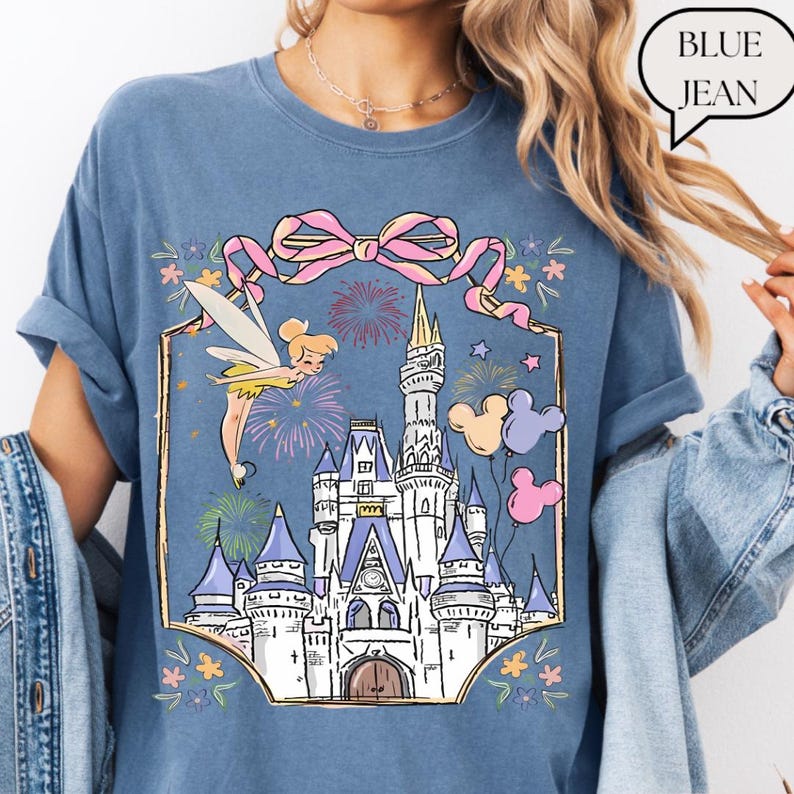 Puede incluir: Camiseta azul vaquero con una ilustraci&oacute;n de castillo con Campanilla, fuegos artificiales y globos de Mickey Mouse. Un lazo rosa y detalles florales enmarcan el dise&ntilde;o del castillo. El texto "BLUE JEAN" est&aacute; en una burbuja.
