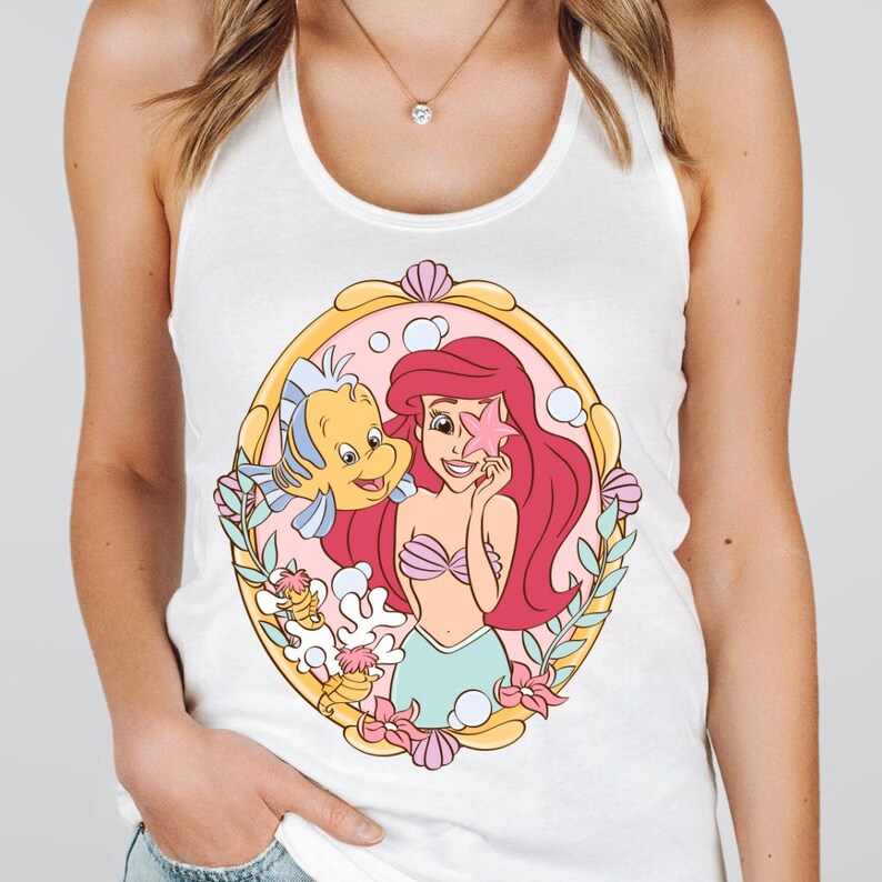 May include: White tank top featuring a colorful illustration of Ariel from The Little Mermaid. Ariel is holding a pink starfish, with Flounder beside her. The design is framed by a gold oval with pink and blue accents.