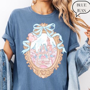 Puede incluir: Camiseta azul vaquero con una ilustraci&oacute;n detallada de un castillo de cuento de hadas dentro de un marco dorado adornado. El marco est&aacute; adornado con un lazo y cintas de color azul claro. El texto "BLUE JEAN" est&aacute; en una burbuja.