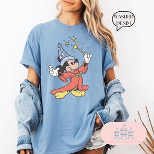 May include: Light blue t-shirt featuring a cartoon Mickey Mouse dressed as a wizard, holding a wand with stars. The shirt has rolled-up sleeves and the text "Washed Denim" in a speech bubble. The shirt is from "SHIRTSEVERAFTERLLC".