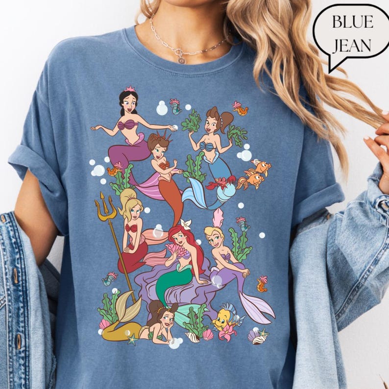 Op de afbeelding: Blauwe spijkerbroekkleurige t-shirt met een kleurrijke illustratie van zeemeerminnen, zeewier en vissen. De afbeelding bevat zeemeerminnen met verschillende haarkleuren en zeemeerminstaarten. De tekst "BLUE JEAN" staat in een tekstballon.