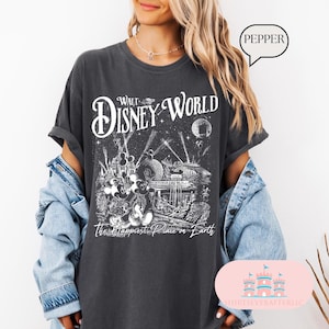 May include: Dark gray t-shirt featuring a Walt Disney World graphic. The design includes the words "Walt Disney World" in a stylized font, along with illustrations of iconic park elements and the phrase "The Happiest Place on Earth."