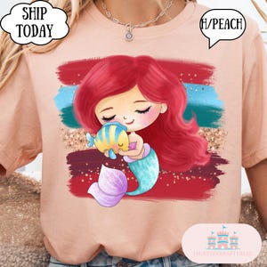 Puede incluir: Camiseta color melocot&oacute;n con una sirena de dibujos animados con pelo rojo, cola turquesa y un pez amarillo y azul. El dise&ntilde;o incluye un fondo rojo, azul y dorado. El texto en la camiseta dice "SHIP TODAY" y "H/PEACH".