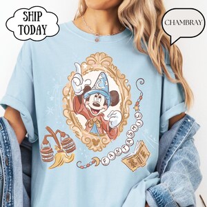 K&ouml;nnte beinhalten: Hellblaues T-Shirt mit einer gerahmten Illustration von Mickey Mouse in einem Zauberer-Outfit. Das Design enth&auml;lt das Wort "FANTASMIC" und andere magische Elemente. Das Shirt hat hochgekrempelte &Auml;rmel und den Text "SHIP TODAY" in einer wolkenf&ouml;rmigen Blase.
