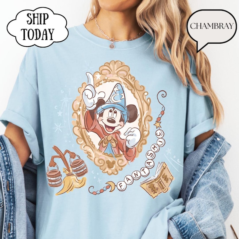K&ouml;nnte beinhalten: Hellblaues T-Shirt mit einer gerahmten Illustration von Mickey Mouse in einem Zauberer-Outfit. Das Design enth&auml;lt das Wort "FANTASMIC" und andere magische Elemente. Das Shirt hat hochgekrempelte &Auml;rmel und den Text "SHIP TODAY" in einer wolkenf&ouml;rmigen Blase.
