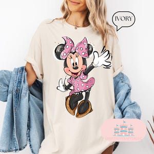 Puede incluir: Una camiseta beige con una imagen de dibujos animados de Minnie Mouse con un vestido rosa de lunares y un lazo rosa. El texto "IVORY" est&aacute; en un bocadillo de di&aacute;logo blanco en la esquina superior derecha de la imagen.