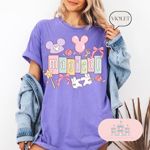 Op de afbeelding: Violetkleurig T-shirt met het woord "magical" in pastelkleuren, met cartoonafbeeldingen van Mickey Mouse-oren, een toverstaf, strikken, rozen en een konijn. Het shirt heeft een relaxte pasvorm en korte mouwen.