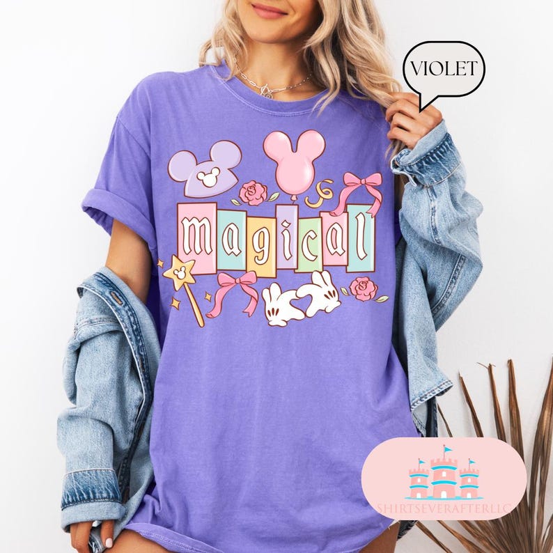 Op de afbeelding: Violetkleurig T-shirt met het woord "magical" in pastelkleuren, met cartoonafbeeldingen van Mickey Mouse-oren, een toverstaf, strikken, rozen en een konijn. Het shirt heeft een relaxte pasvorm en korte mouwen.