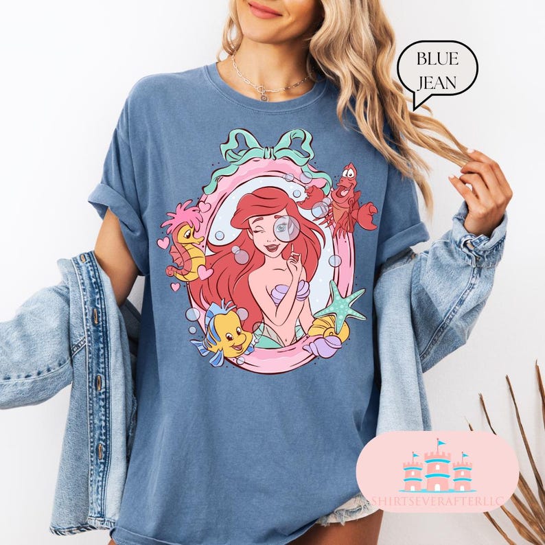 K&ouml;nnte beinhalten: Blaues Jeans-T-Shirt mit einer Cartoon-Illustration von Arielle, der Meerjungfrau. Das Design zeigt Arielle, Fabius, Sebastian und andere Meereskreaturen in einem rosa und wei&szlig;en Rahmen. Der Text "BLUE JEAN" befindet sich in einer Sprechblase.
