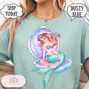 May include: A dusty blue t-shirt with a watercolour illustration of a mermaid in a seashell. The mermaid has red hair, a purple seashell bra, and a teal tail. The text "SHIP TODAY" and "DUSTY BLUE" are in speech bubbles.