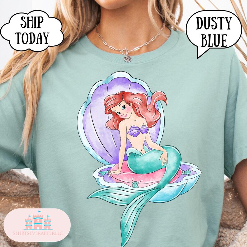 May include: A dusty blue t-shirt with a watercolour illustration of a mermaid in a seashell. The mermaid has red hair, a purple seashell bra, and a teal tail. The text "SHIP TODAY" and "DUSTY BLUE" are in speech bubbles.