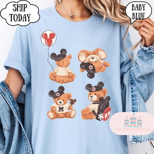 May include: A light blue t-shirt featuring a graphic print of four brown teddy bears wearing black mouse-ear hats. One bear holds a red balloon, another wears a white shirt with the letter M, and one carries a backpack with red and white details.