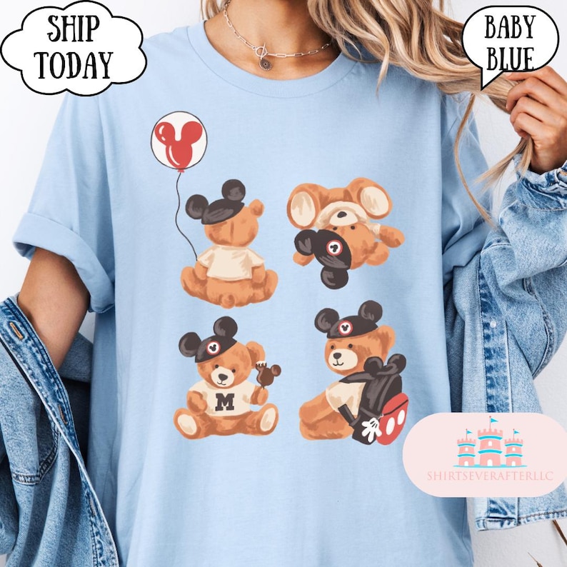 May include: A light blue t-shirt featuring a graphic print of four brown teddy bears wearing black mouse-ear hats. One bear holds a red balloon, another wears a white shirt with the letter M, and one carries a backpack with red and white details.
