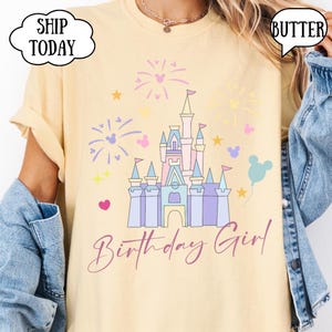May include: A pale yellow t-shirt with a castle graphic, fireworks, stars, and a balloon. The text "Birthday Girl" is in cursive. The shirt has the word "Butter" in a speech bubble. The shirt is a light yellow color.