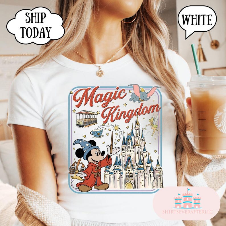 May include: White t-shirt featuring a colorful graphic of Mickey Mouse in a sorcerer's outfit, a castle, and the words "Magic Kingdom." The design includes a trolley car, Dumbo, and Tinkerbell. The shirt has a round neck.