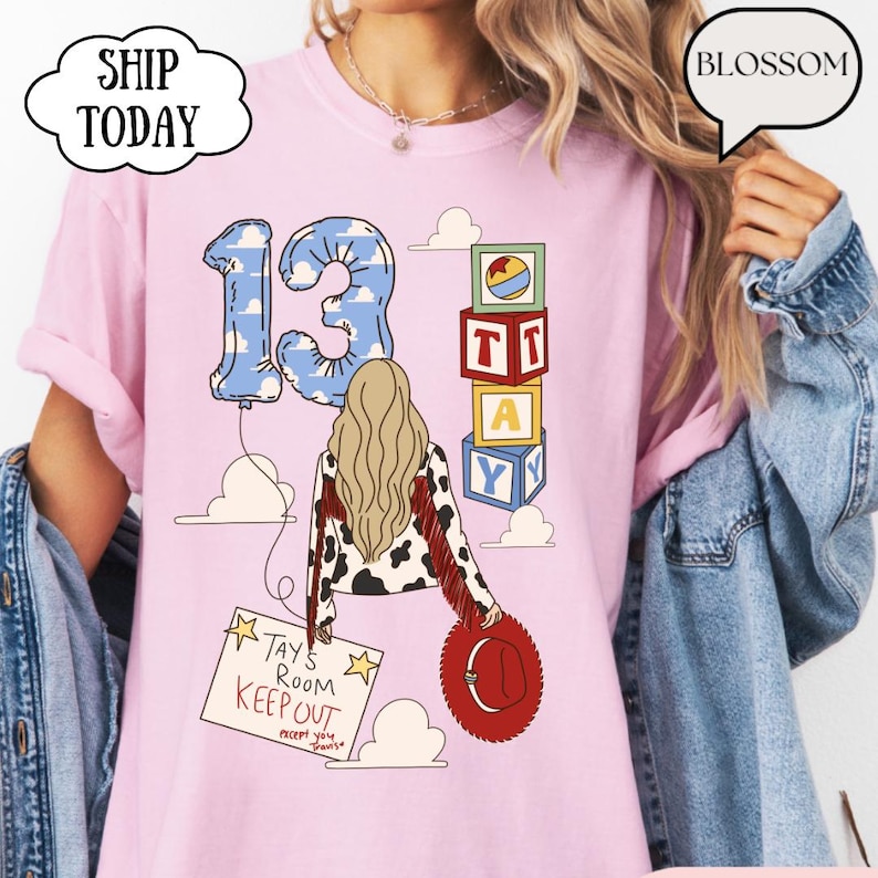 May include: A light pink t-shirt featuring a graphic of a person with long blonde hair wearing a cow-print jacket with red fringe. The design includes a blue cloud-patterned number 13 balloon, alphabet blocks spelling TAY, and a sign reading TAY'S ROOM KEEP OUT.