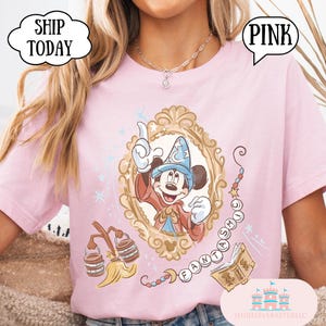 K&ouml;nnte beinhalten: Hellrosa T-Shirt mit einer gerahmten Illustration von Mickey Mouse in einem Zauberer-Outfit. Das Design enth&auml;lt das Wort "FANTASMIC" und andere magische Elemente. Das Shirt hat die Texte "PINK" und "SHIP TODAY" in Sprechblasen.