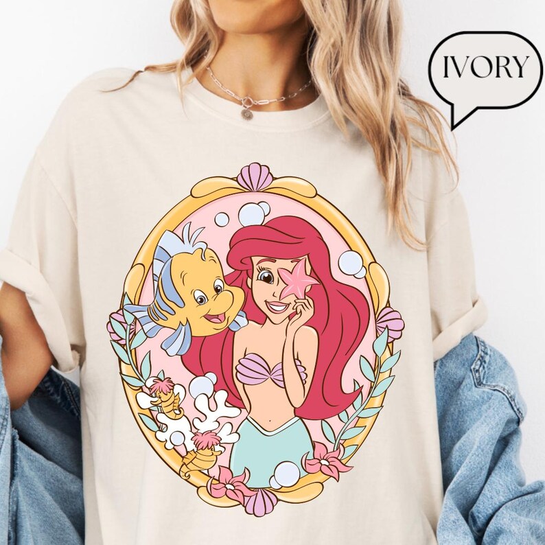 May include: Ivory-colored t-shirt featuring a colorful graphic of a mermaid holding a starfish, with a yellow fish and floral accents. The design is framed in a gold oval. The word "IVORY" is in a speech bubble.