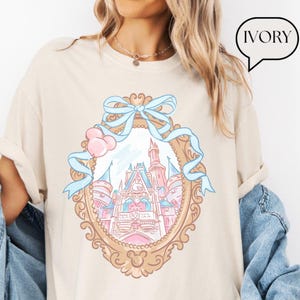 Puede incluir: Camiseta color marfil con un dise&ntilde;o de castillo caprichoso dentro de un marco ornamentado, adornado con una cinta azul claro y globos rosas. El castillo est&aacute; representado en tonos pastel rosas y azules. La palabra "IVORY" est&aacute; en una burbuja.