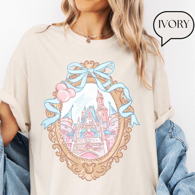 Puede incluir: Camiseta color marfil con un dise&ntilde;o de castillo caprichoso dentro de un marco ornamentado, adornado con una cinta azul claro y globos rosas. El castillo est&aacute; representado en tonos pastel rosas y azules. La palabra "IVORY" est&aacute; en una burbuja.