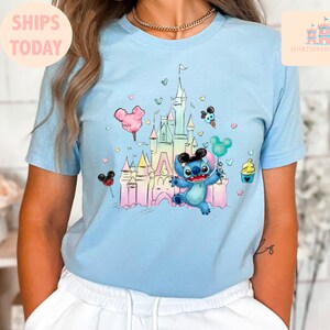 May include: A light blue t-shirt with a colorful illustration of a cartoon Stitch character holding a balloon in front of a pastel-colored castle. The castle is surrounded by hearts and other cartoon details.