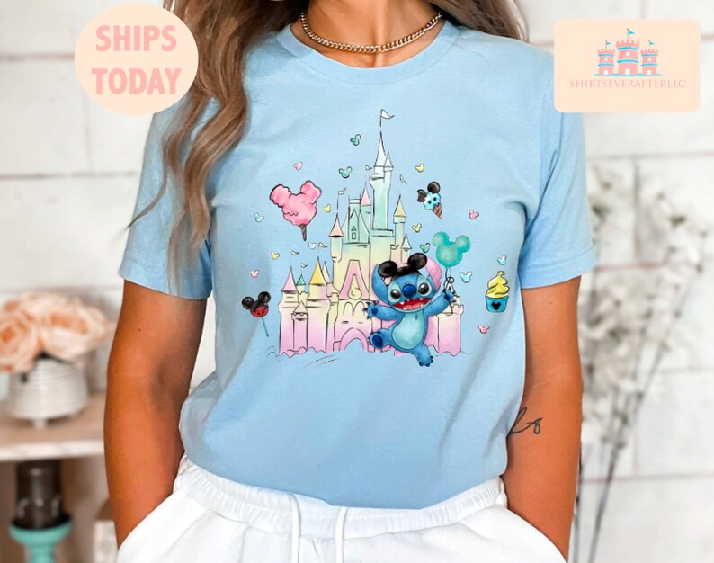 May include: A light blue t-shirt with a colorful illustration of a cartoon Stitch character holding a balloon in front of a pastel-colored castle. The castle is surrounded by hearts and other cartoon details.