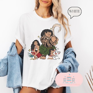 May include: White t-shirt with a graphic print of Maui, Moana, and Pua from the Disney movie Moana. The characters are depicted in a cartoon style with vibrant colors.