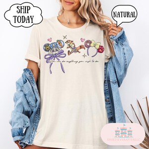 May include: Cream-colored t-shirt featuring cartoon character designs and the text "You can be anything you wish to be." The shirt has a relaxed fit and is paired with a denim jacket.