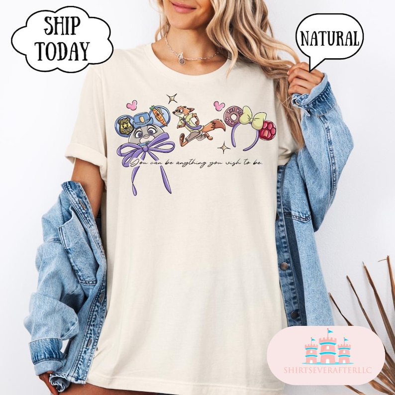 May include: Cream-colored t-shirt featuring cartoon character designs and the text "You can be anything you wish to be." The shirt has a relaxed fit and is paired with a denim jacket.