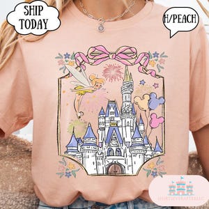 Puede incluir: Camiseta color melocot&oacute;n con una ilustraci&oacute;n de un castillo de cuento de hadas, fuegos artificiales y un hada. El dise&ntilde;o incluye un lazo rosa, globos de Mickey Mouse y detalles florales. El texto "SHIP TODAY" es visible.
