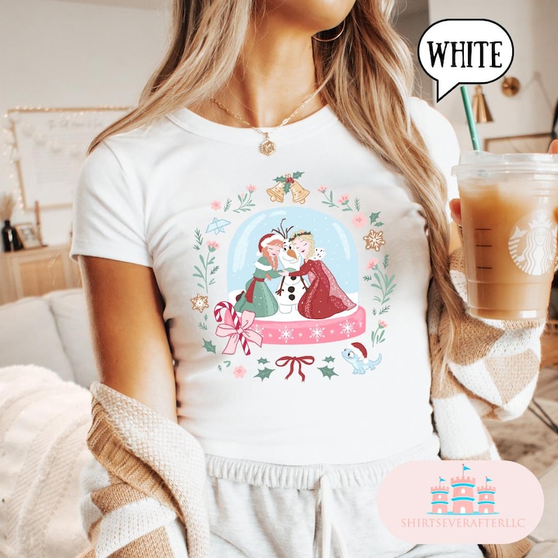 May include: White t-shirt featuring a snow globe design with two characters, a snowman, and holiday elements. The snow globe is surrounded by a wreath of leaves, flowers, and bells. The word "WHITE" is in a speech bubble.