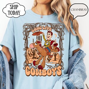 May include: Light blue t-shirt featuring a cartoon cowboy riding a horse, with the words "Long Live Cowboys" in a vintage style. The design includes a cowboy character and a horse in a desert landscape.