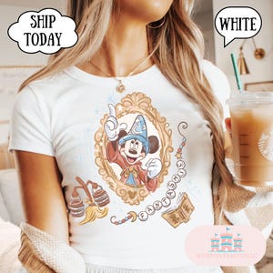 K&ouml;nnte beinhalten: Wei&szlig;es T-Shirt mit einem Cartoon-Mickey-Mouse-Zauberer in einem goldenen Rahmen. Das Shirt enth&auml;lt das Wort "FANTASMIC" und andere Grafiken. Der Text "SHIP TODAY" und "WHITE" sind ebenfalls sichtbar.