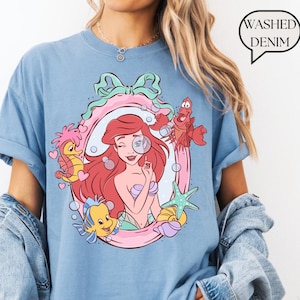 May include: Light blue t-shirt featuring a colorful illustration of a mermaid with red hair, holding a magnifying glass, surrounded by sea creatures. The design is framed in a pink and green oval. The text "Washed Denim" is in a speech bubble.