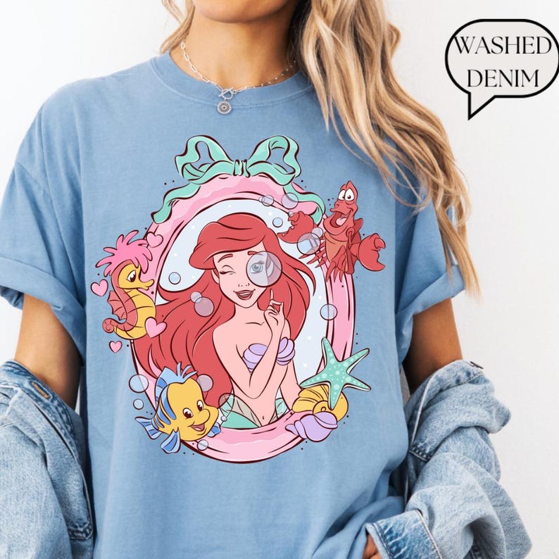 May include: Light blue t-shirt featuring a colorful illustration of a mermaid with red hair, holding a magnifying glass, surrounded by sea creatures. The design is framed in a pink and green oval. The text "Washed Denim" is in a speech bubble.