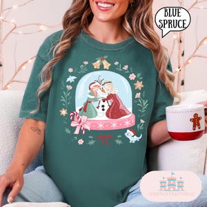 Puede incluir: Camiseta de color verde azulado con un diseño de globo de nieve con personajes de dibujos animados, copos de nieve y detalles florales. El texto "BLUE SPRUCE" está en un círculo blanco. Se ve una taza blanca con un hombre de jengibre.
