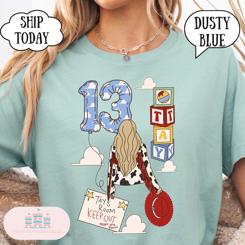 May include: A dusty blue t-shirt featuring a graphic of a person with long blonde hair, a cow-print jacket with red fringe, and a red hat. The design includes a balloon shaped like the number 13, alphabet blocks, and a sign reading TAYS ROOM KEEP OUT.
