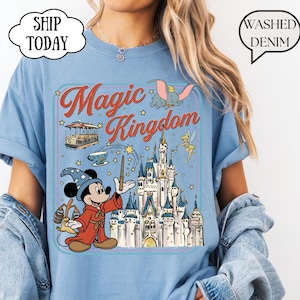 May include: Light blue t-shirt with a graphic of Mickey Mouse as a wizard, a castle, and the words "Magic Kingdom." The design includes Dumbo, Tinkerbell, and a trolley car. The shirt has a relaxed fit and short sleeves.