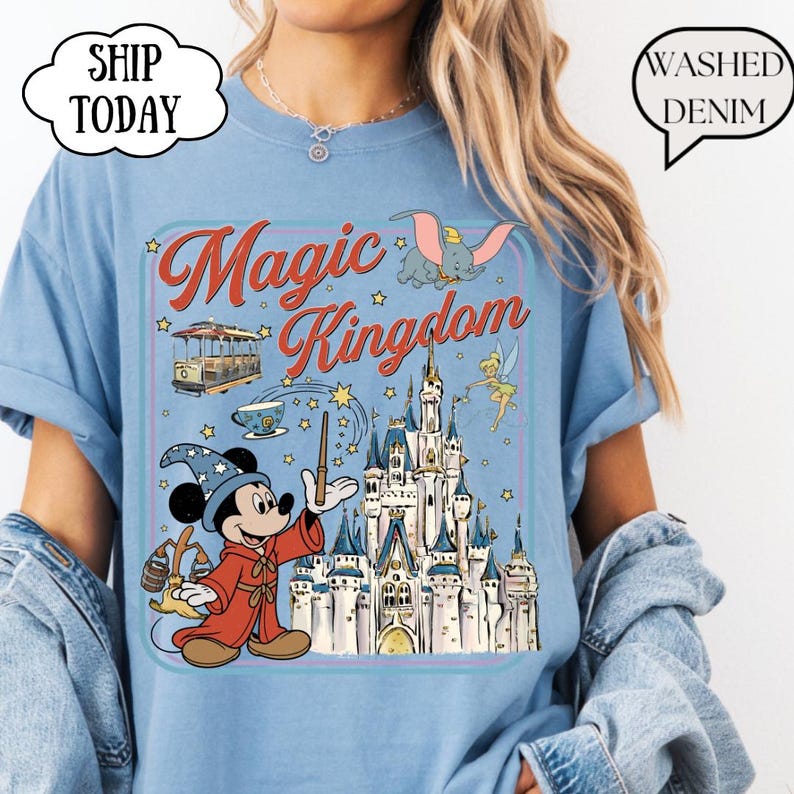 May include: Light blue t-shirt with a graphic of Mickey Mouse as a wizard, a castle, and the words "Magic Kingdom." The design includes Dumbo, Tinkerbell, and a trolley car. The shirt has a relaxed fit and short sleeves.