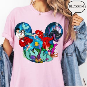 Puede incluir: Camiseta rosa claro con un dise&ntilde;o de personajes de La Sirenita, dispuestos en una silueta de Mickey Mouse. El dise&ntilde;o incluye a Ariel, el Pr&iacute;ncipe Eric, Flounder y Ursula. La palabra "BLOSSOM" est&aacute; en una burbuja.