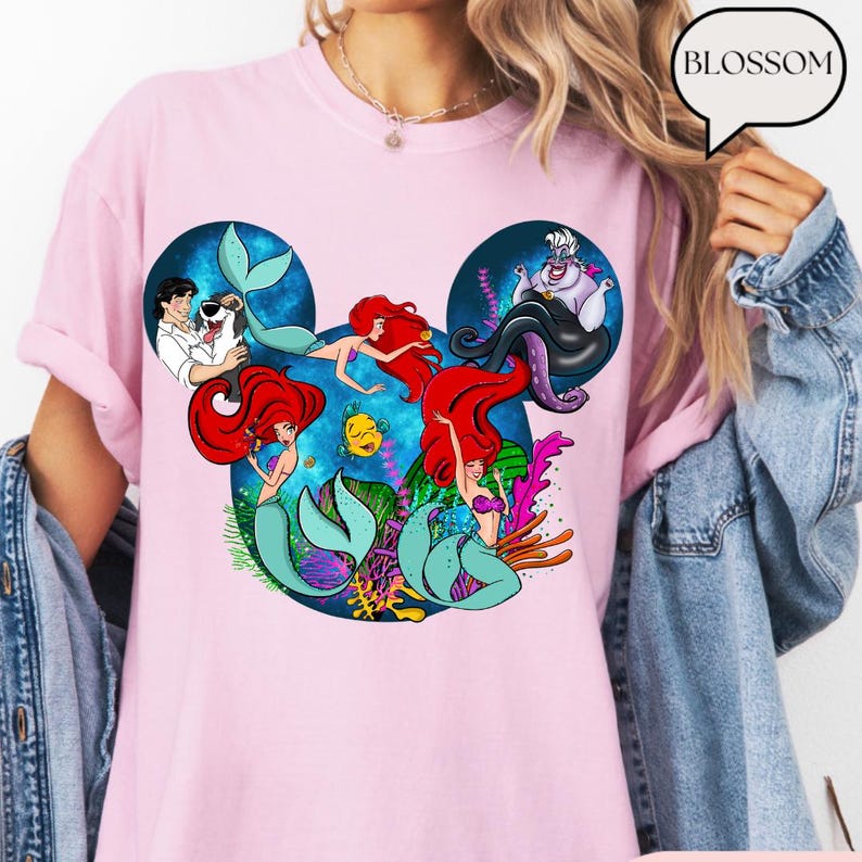 Puede incluir: Camiseta rosa claro con un dise&ntilde;o de personajes de La Sirenita, dispuestos en una silueta de Mickey Mouse. El dise&ntilde;o incluye a Ariel, el Pr&iacute;ncipe Eric, Flounder y Ursula. La palabra "BLOSSOM" est&aacute; en una burbuja.