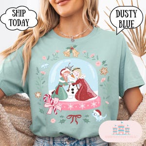 Puede incluir: Camiseta azul polvoriento con un diseño de globo de nieve con dos personajes de dibujos animados, un muñeco de nieve y detalles florales. La camiseta tiene los textos "SHIP TODAY" y "DUSTY BLUE" en bocadillos. También es visible la marca "SHIRTSEVERAFTERLLC".