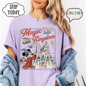 May include: A light purple t-shirt featuring a graphic of Mickey Mouse as a sorcerer in front of a castle, with the words "Magic Kingdom". The design includes Dumbo, Tinkerbell, and a cable car.